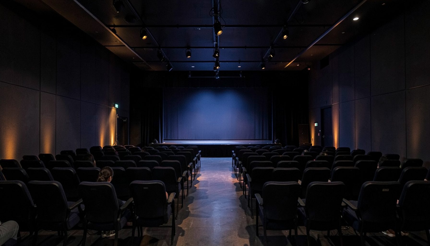 Dark conference hall with rows of chairs facing a softly lit stage, deep navy and charcoal tones with warm amber accent lighting